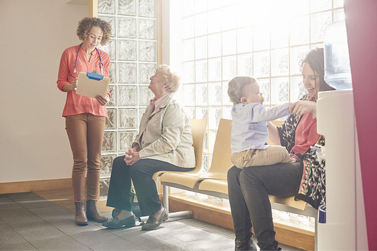 Waiting room with four patients - a mother playing with her child sitting down, and a young lady medical professional talking with a seated patient waiting in the room
